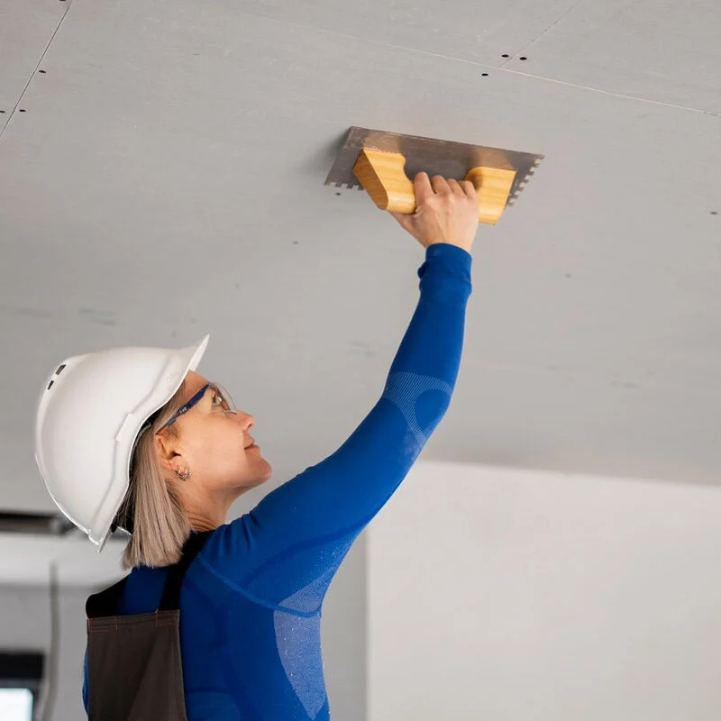 A woman in a hard hat is seen holding a piece of wood, indicating her involvement in construction or woodworking activities.